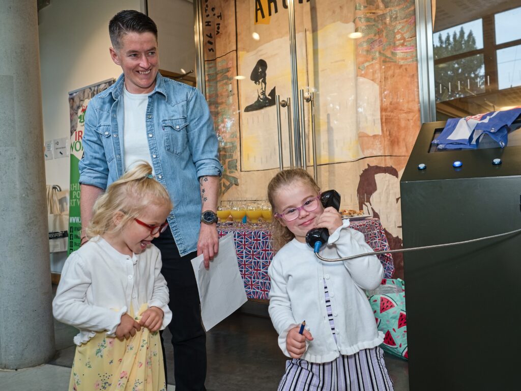A family enjoying a visit to the museum during an exhibition launch event for Children and Military Lives. Two young girls use a gallery interactive, one holding a telephone to their ear, while their parent looks on smiling
