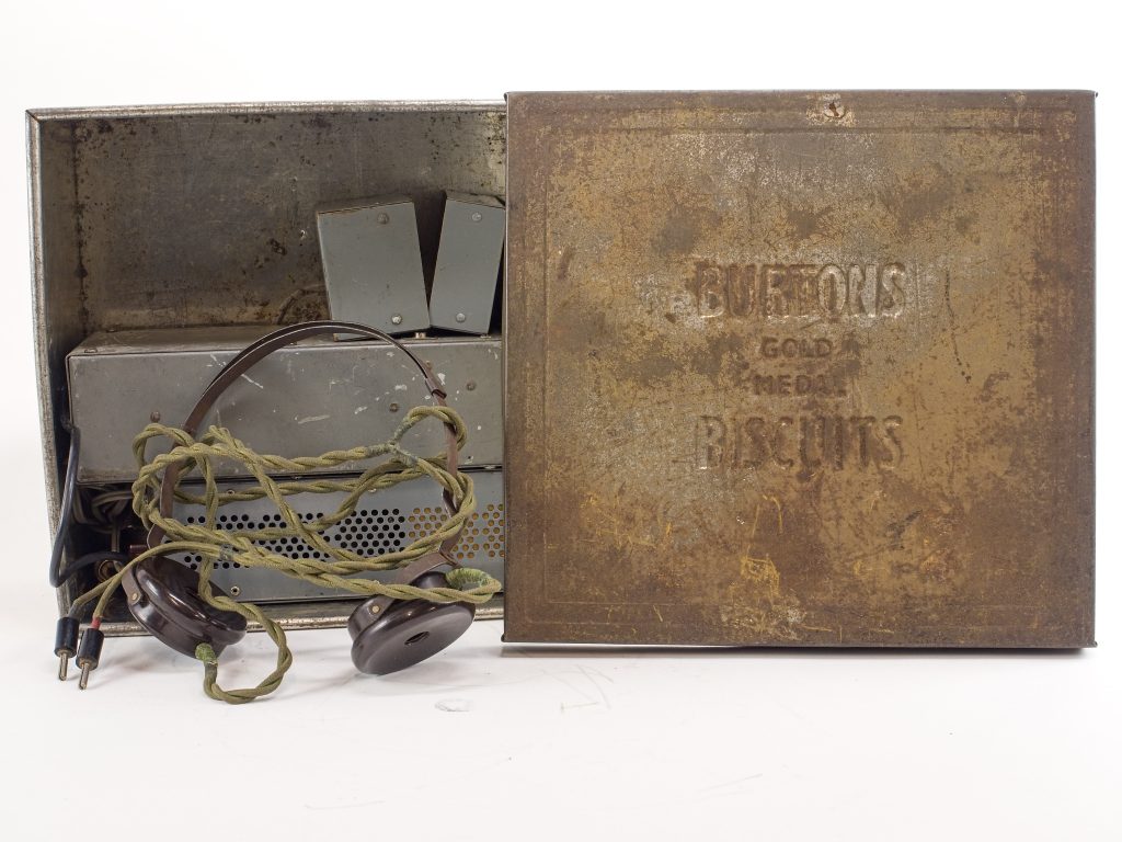 Square steel biscuit tin stands on its side with the lid removed and standing beside it. The tin is largely rusted with age, little steel shines through. Nestled inside the tin is a set of radio equipment and a headset, used by SOE agents during the Second World War to listen in on radio transmissions.