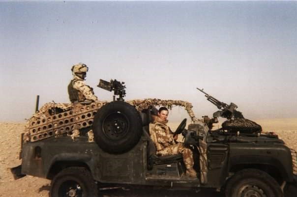 Jessica Lakin sits behind the wheel of an Army Land Rover. She wars camouflaged combat fatigues, while another soldier stands on the back of the vehicle manning a machine gun. Desert sands and a hazy blue sky can be seen in the background.