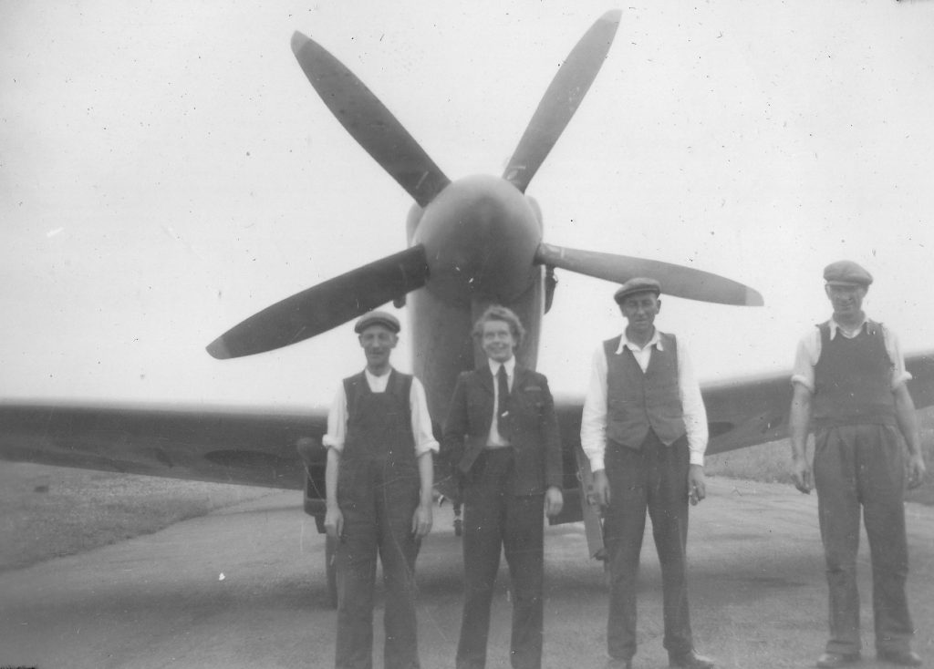 A black and white photograph. Mary Wilkins, in an ATA jacket and necktie, stands in front of the nose of a propellor aircraft, facing the camera. Three men in flat caps, shirts and waistcoats stand beside her, one to her left, two to her right to her right.