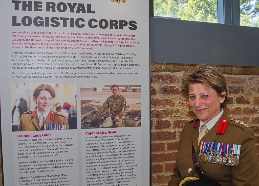 Colonel Lucy Giles stands next to an exhibition panel which features images and text telling the story of women in the Royal Logistic Corps in recent years. She is smiling at the camera, and is wearing her military uniform with large set of medals pinned to her chest. Her arms are crossed in front of her, holding a peaked cap and gloves.