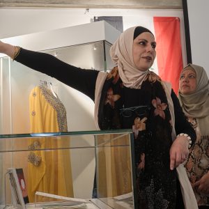 Rana Ibrahim introduces an IWAW exhibition. She has an arm raised, pointing our displays on a wall behind here. Display cabinets around her contain dresses and artwork by Iraqi women.